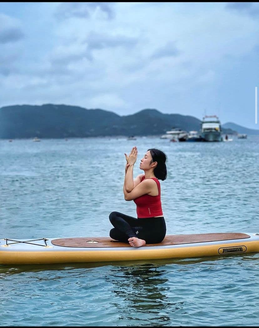 一名女子穿着红色背心在海上的桨板上练习瑜伽,背景是连绵的山脉。