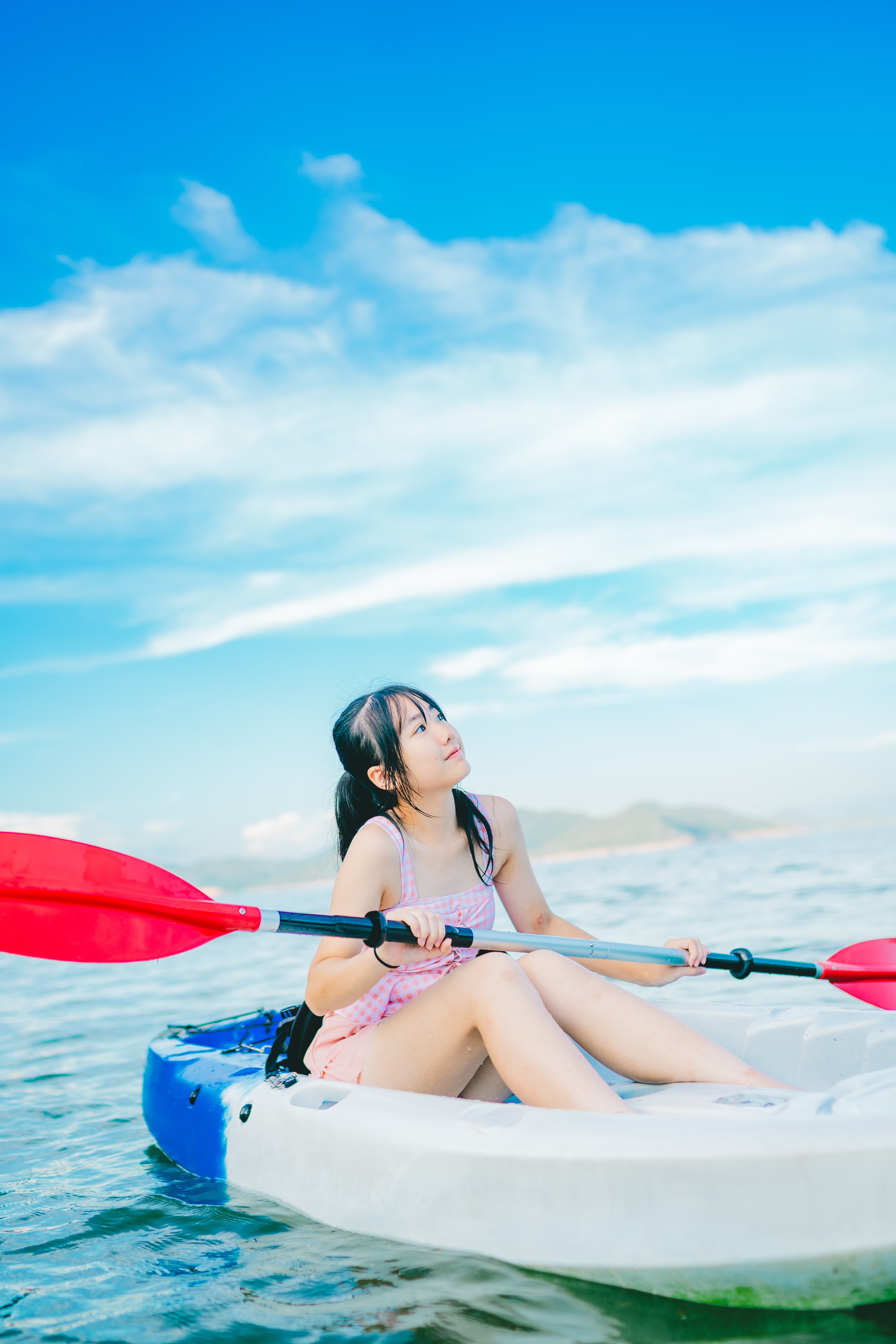 蓝天白云下,一名穿着粉色格纹泳衣的少女在海面上划着皮划艇。
