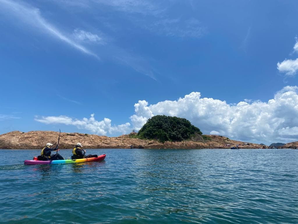 两人在蔚蓝海面上划着彩虹色皮划艇,背景是绿树覆盖的岩石岛屿。